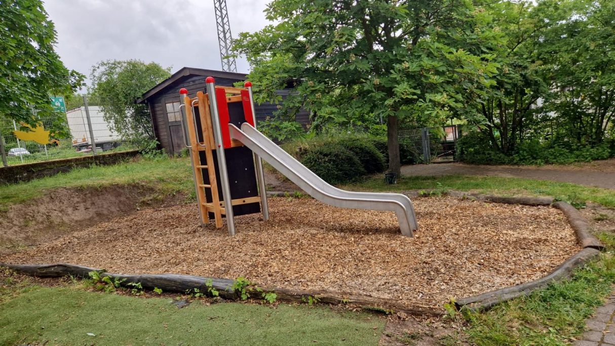 Rutsche auf einem Spielplatz, umgeben von einem holzigen Untergrund und Bäumen.
