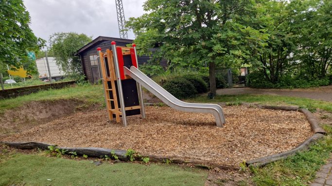 Rutsche auf einem Spielplatz, umgeben von Kies und Bäumen.
