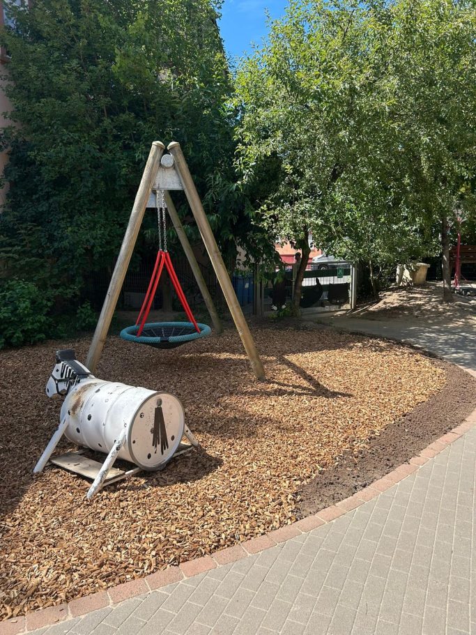 Spielplatz mit einer Schaukel und einer umgestürzten Holztonne, umgeben von Bäumen.