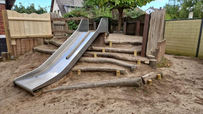 Rutsche aus Metall auf Holzspielplatz mit Holzstufen und sandigem Untergrund.